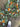 A top-down view of a large autumn wreath decorated with green foliage, pinecones, orange dried pumpkins, and dried orange slices, laid on a stone floor next to a person's white sneaker.