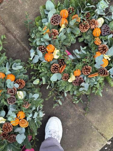 A top-down view of a large autumn wreath decorated with green foliage, pinecones, orange dried pumpkins, and dried orange slices, laid on a stone floor next to a person's white sneaker.